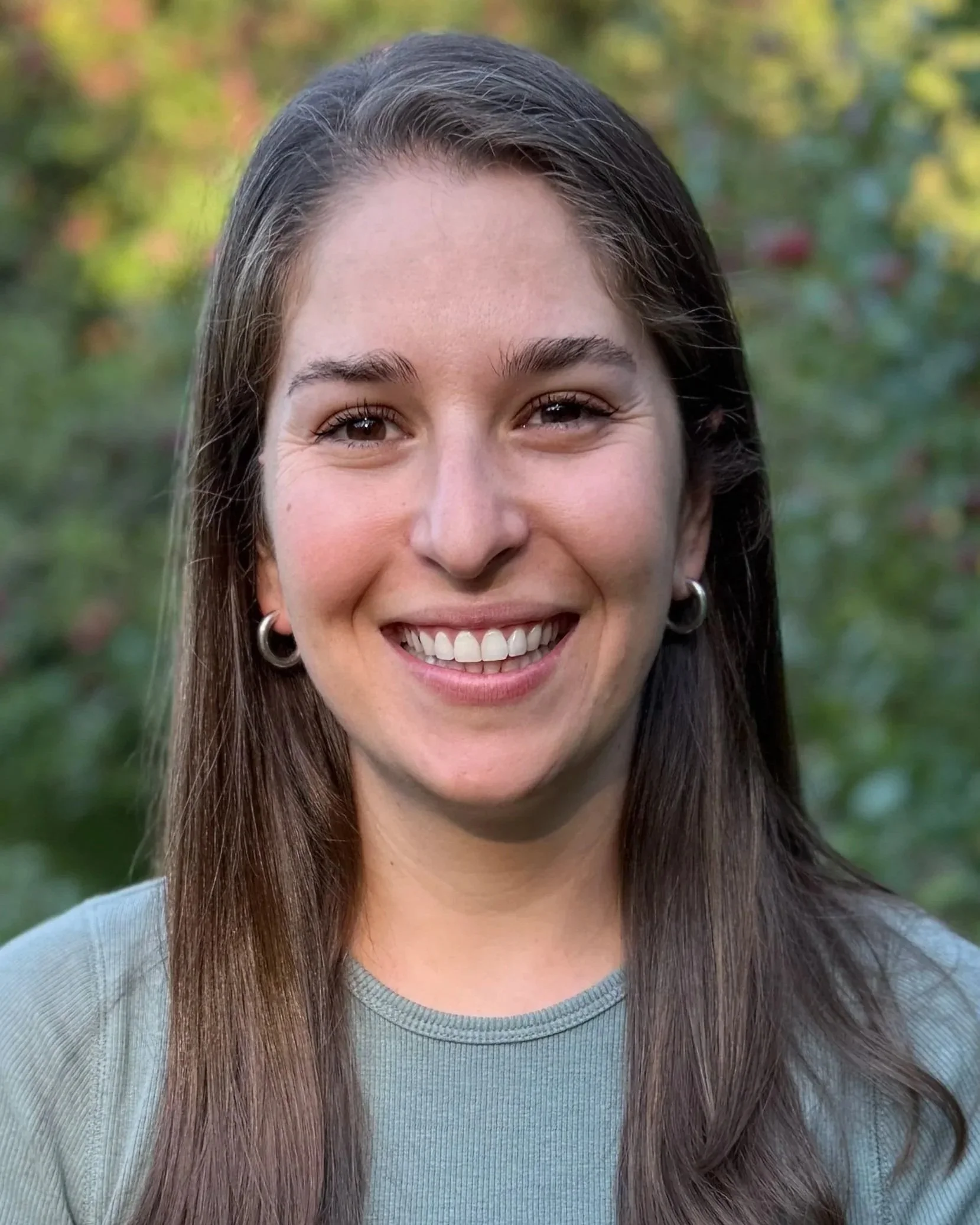 Close-up of a smiling woman with long brown hair, wearing a light blue shirt, standing outdoors with blurred green foliage in the background.