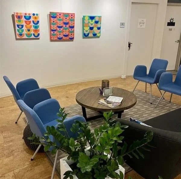 A waiting room with six blue chairs, a round wooden coffee table with magazines and a card, a green plant in the foreground, an abstract colorful artwork on the wall, and a door in the background.