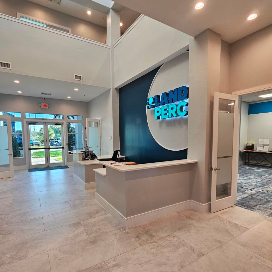 Reception area of a modern office with a white counter, computers, and a blue accent wall with a circular logo that says "LAND PERC" in glowing blue letters. Glass doors and windows reveal a parking lot outside.