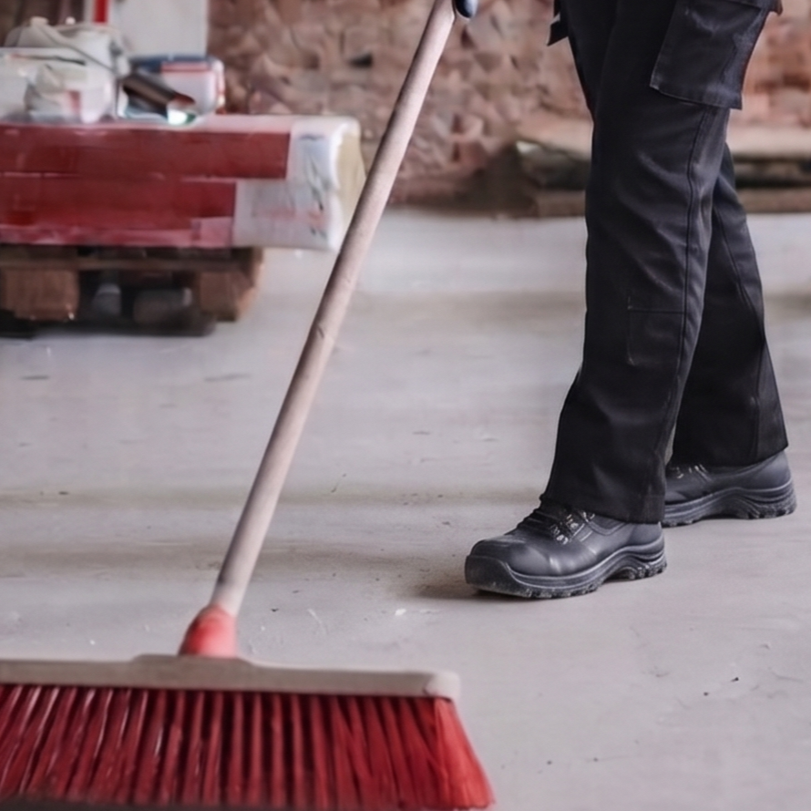 Part of a person wearing black work pants and sturdy black work boots sweeping a concrete floor with a red push broom in a workshop or construction site.