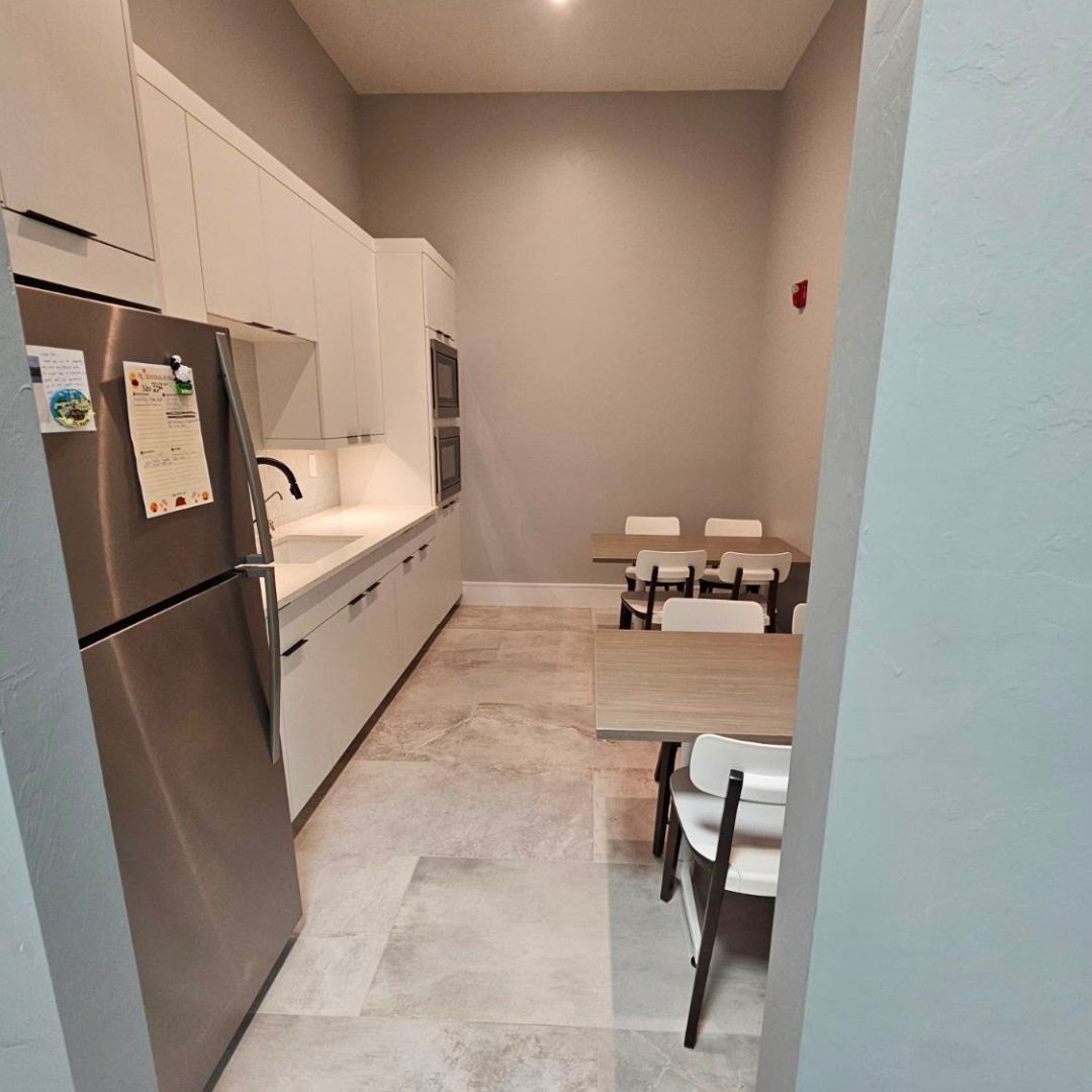 A small kitchen with white cabinets, a stainless steel refrigerator, and two built-in ovens. There are two tables with chairs in the corner.