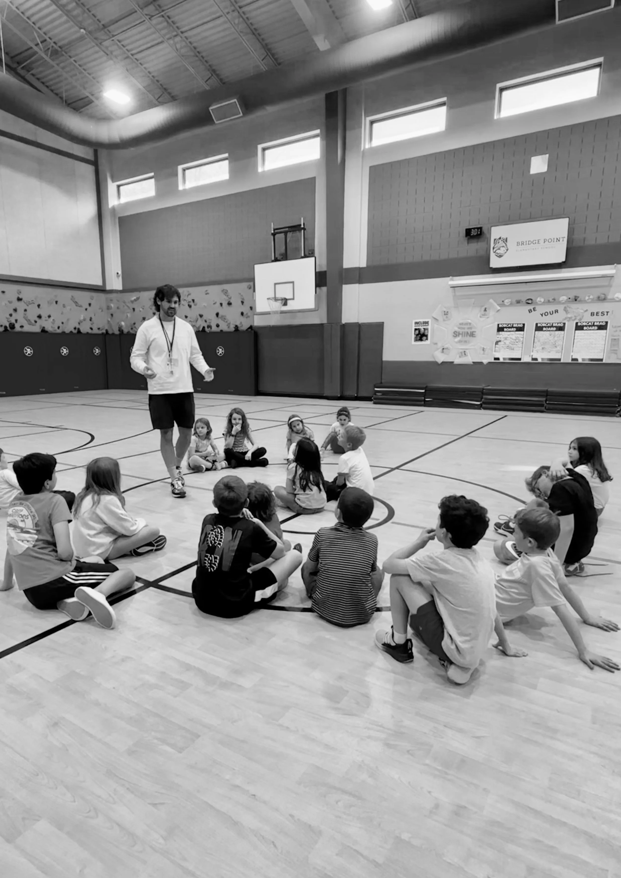 A man is standing and talking to a group of children seated on the gymnasium floor, with a basketball hoop and gym wall in the background.