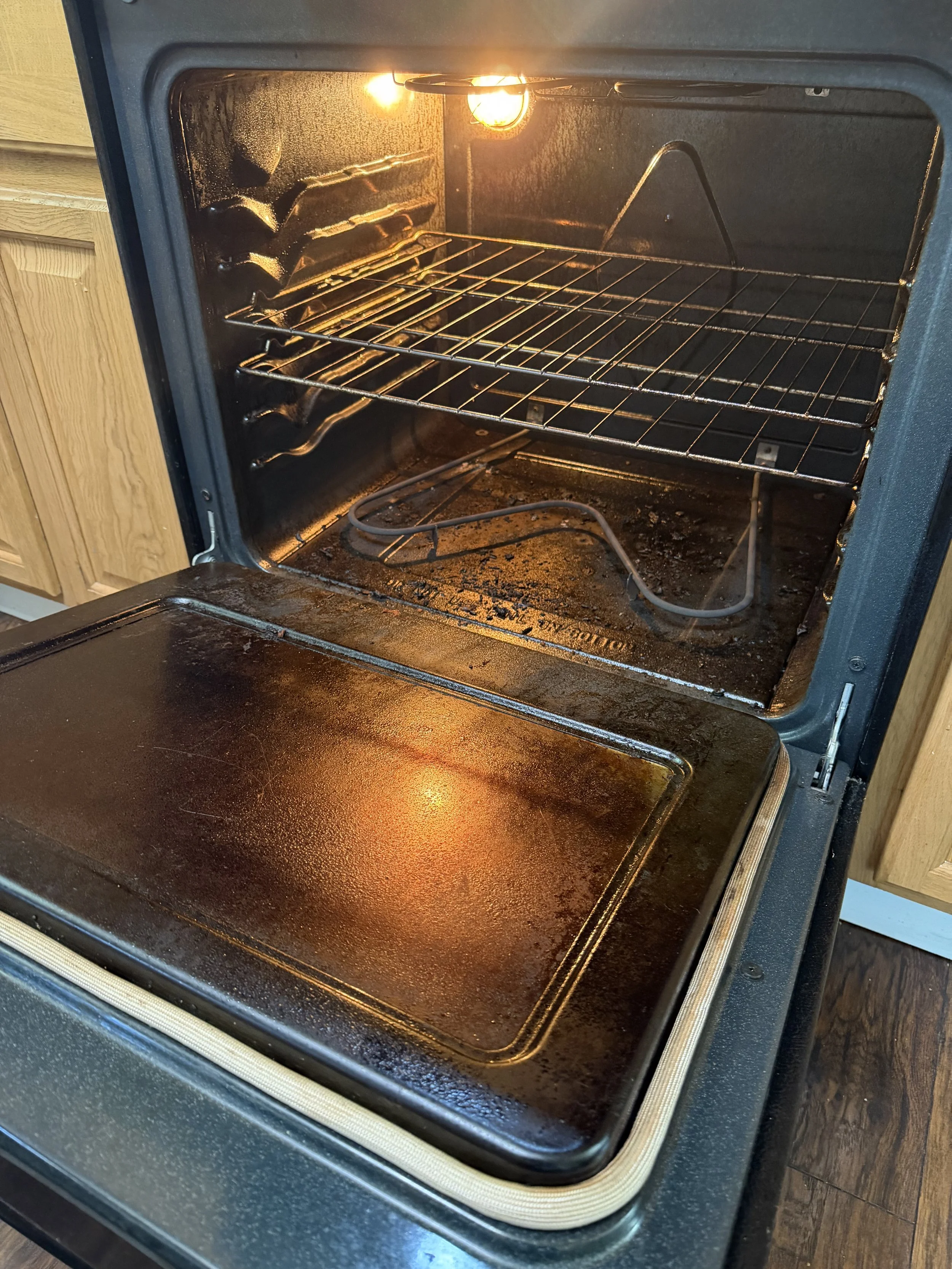 Inside of an oven with two heating elements, one at the top and one at the bottom, and a heated wire rack in the middle. The oven light is on, illuminating the interior which has some soot and residue.