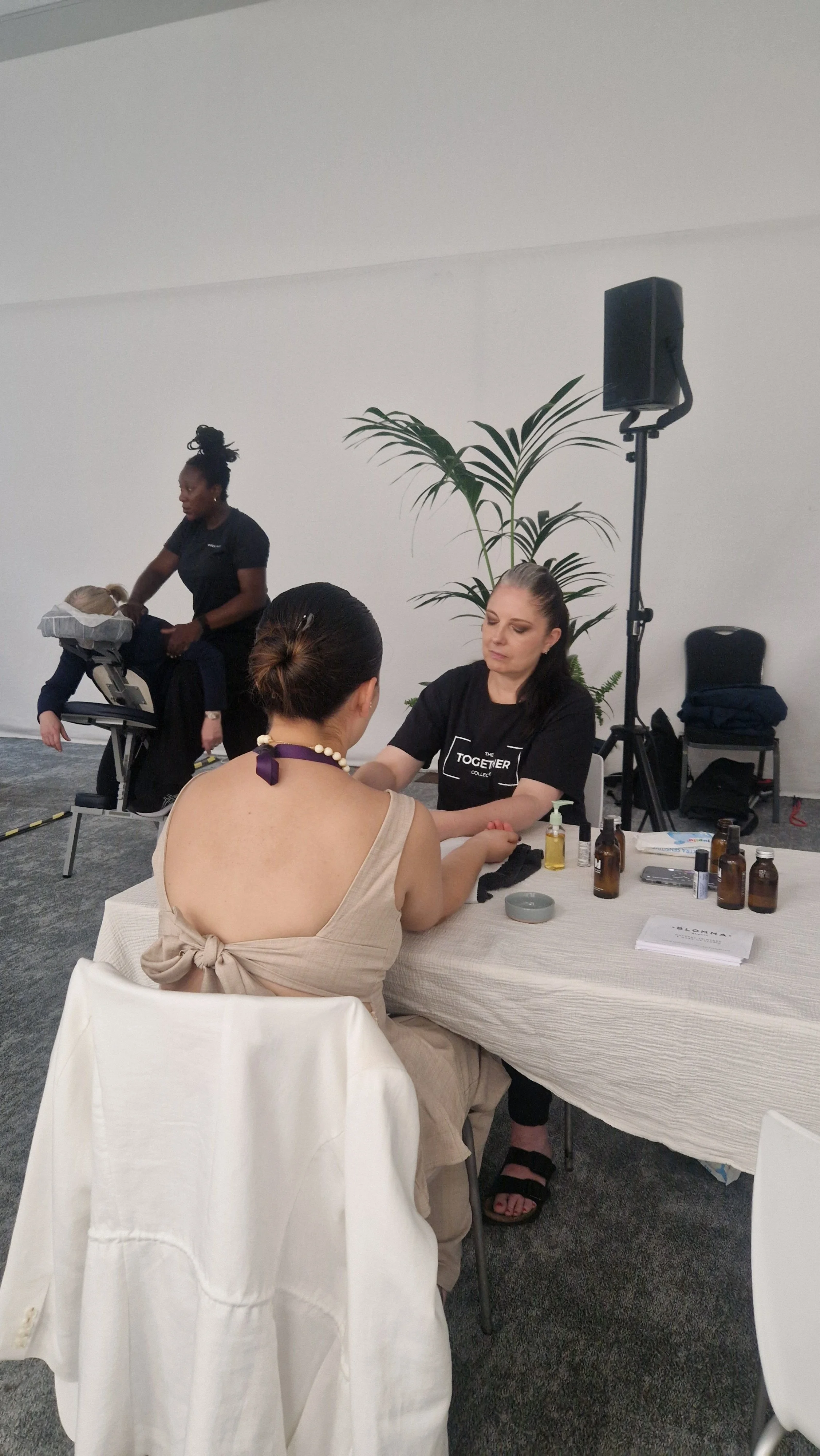 A woman is receiving a facial treatment or skin consultation from a professional seated at a table with skincare products and documents, while another person in the background is lying face down on a massage or therapy table with a practitioner attending to them.