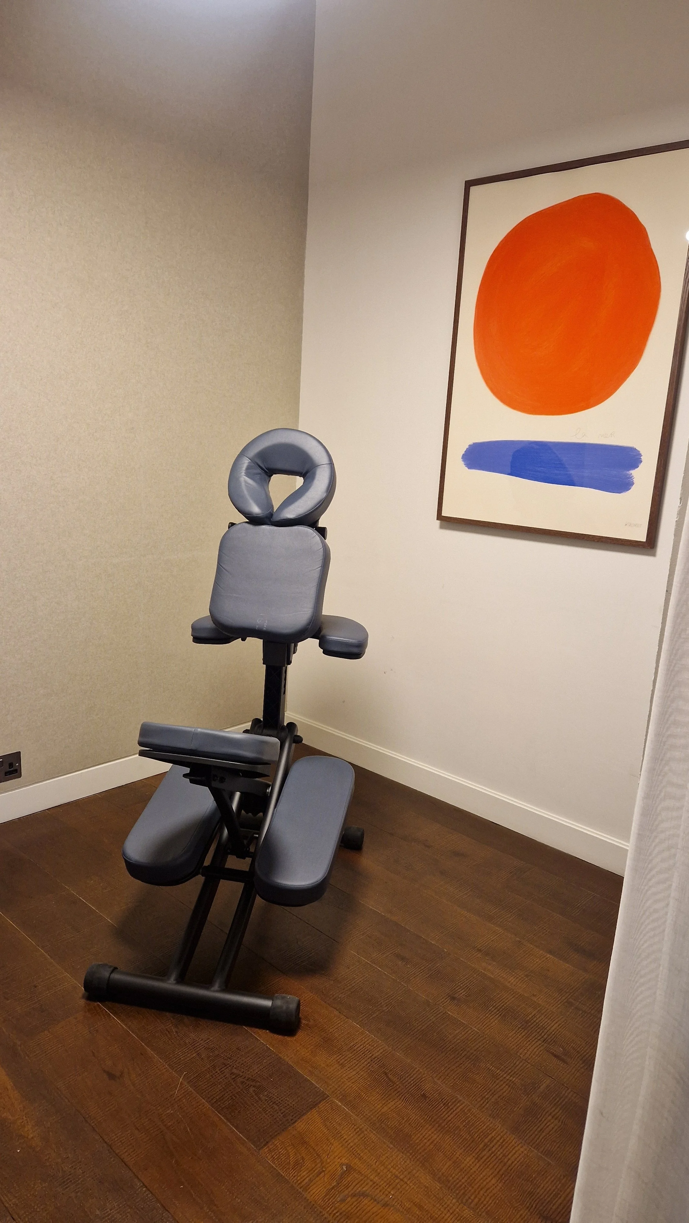 A medical or massage table with a face cradle in a room with wooden flooring, beige and white walls, and a colorful abstract painting with a large orange circle and a blue brush stroke on the wall.