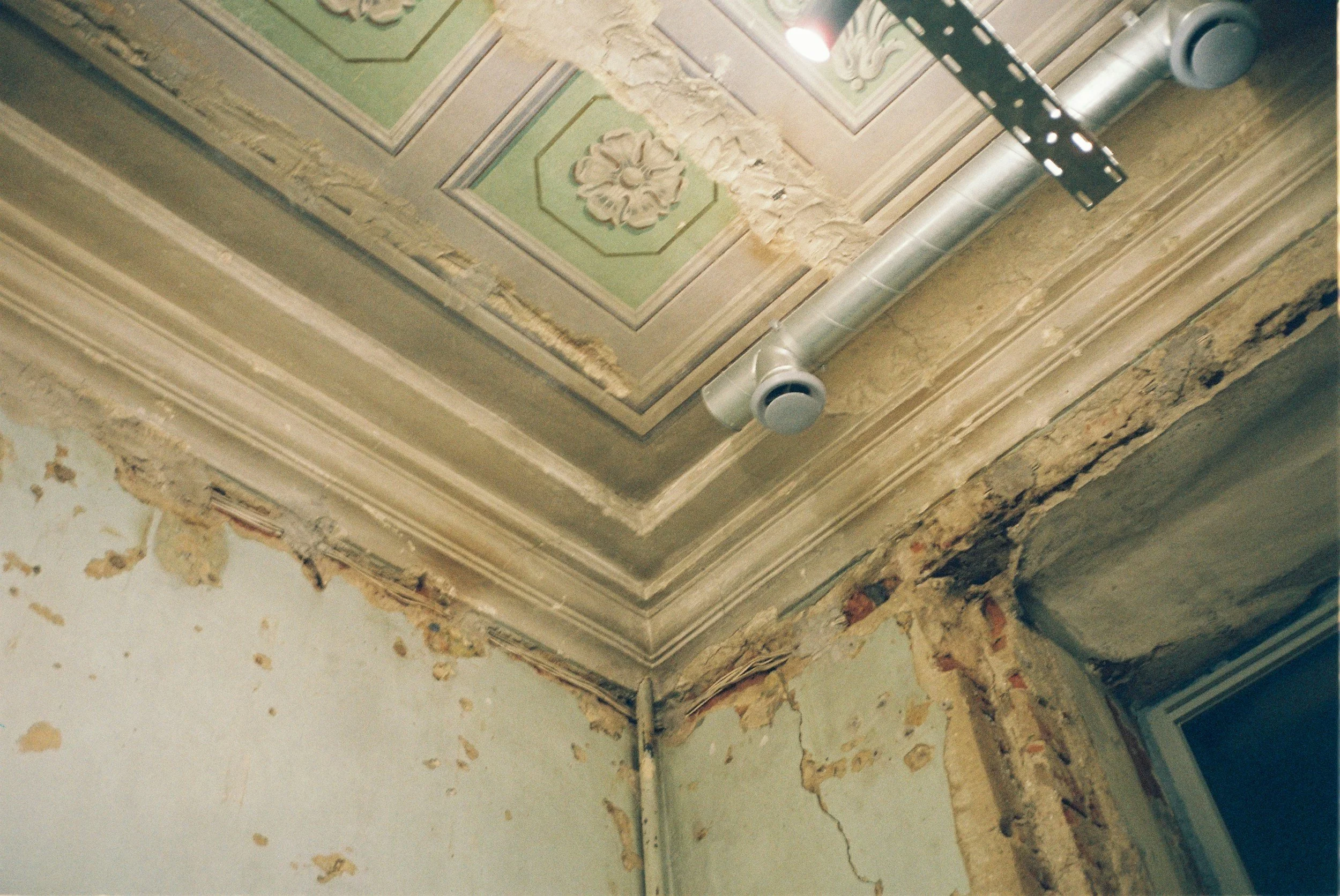 Ceiling with decorative molding undergoing renovations, with peeling paint and exposed walls, and construction equipment attached.