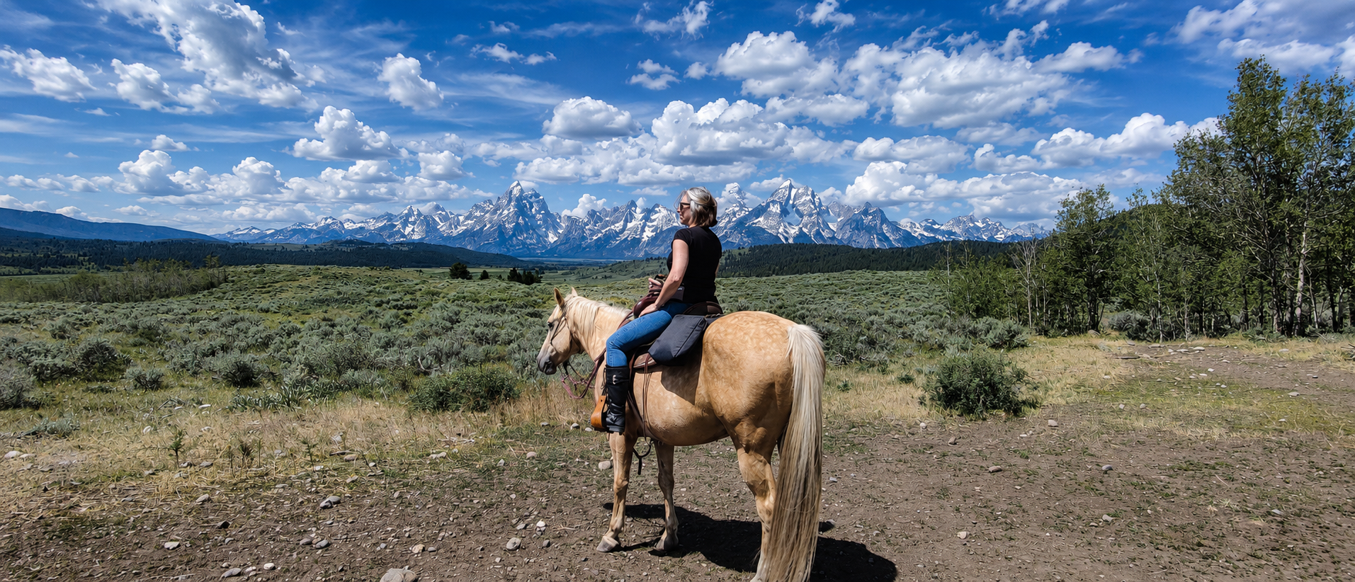 A woman riding a light-colored horse in a mountainous landscape with snow-capped peaks, green trees, and a bright blue sky with clouds.