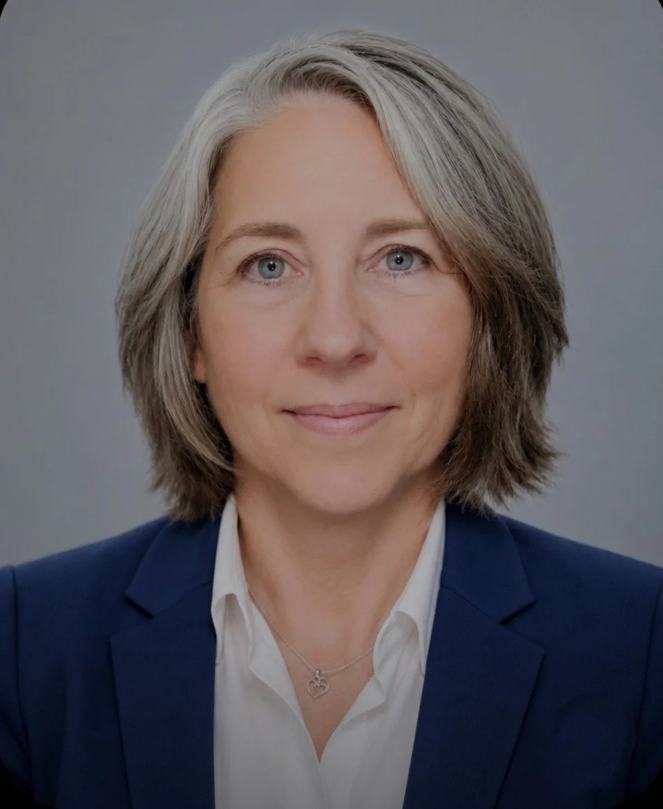 Headshot of a middle-aged woman with gray hair, wearing a navy blazer and white blouse, smiling against a light gray background.