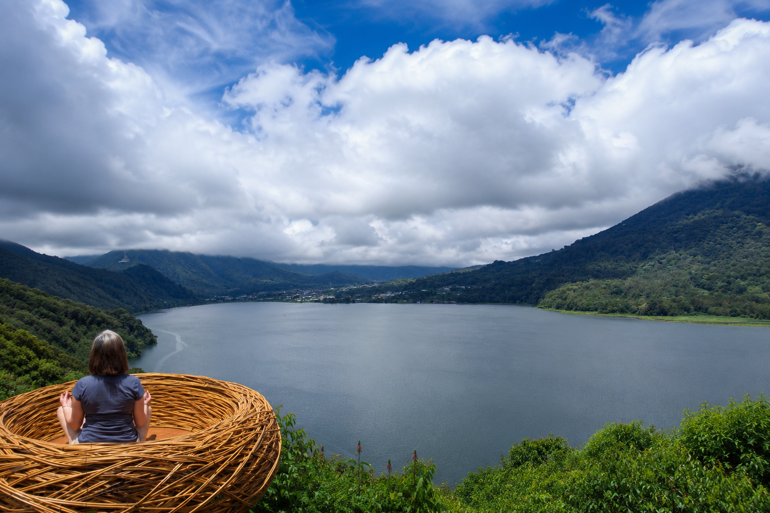 A young girl sits in a wicker nest-shaped chair on a hill overlooking a lake surrounded by green mountains on a cloudy day.