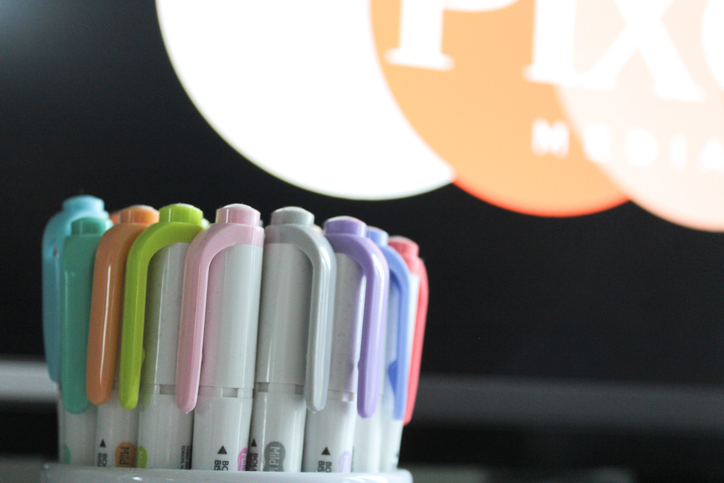 A row of colorful dry erase markers standing upright on a shelf with a background of glowing orange paper lanterns.