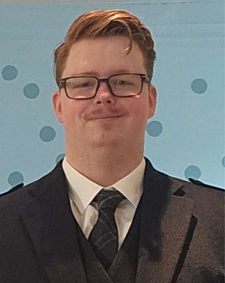 A young man with light skin, glasses, and short reddish-brown hair, wearing a suit with a tie, smiling in front of a light blue background with darker blue polka dots.