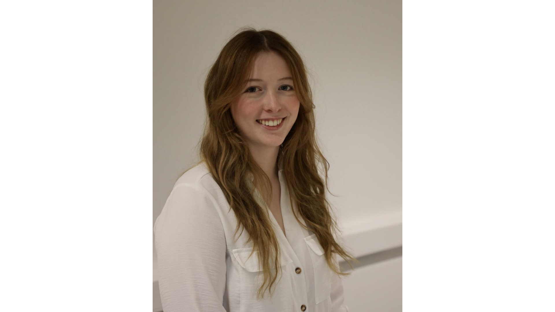 A young woman with long, wavy, auburn hair smiling at the camera against a plain light-colored wall.