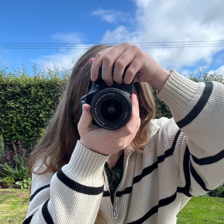 Person with long hair taking a photo in a garden, holding a camera up to their face, wearing a cream-colored sweater with black stripes, with a blue sky and green shrubbery in the background.