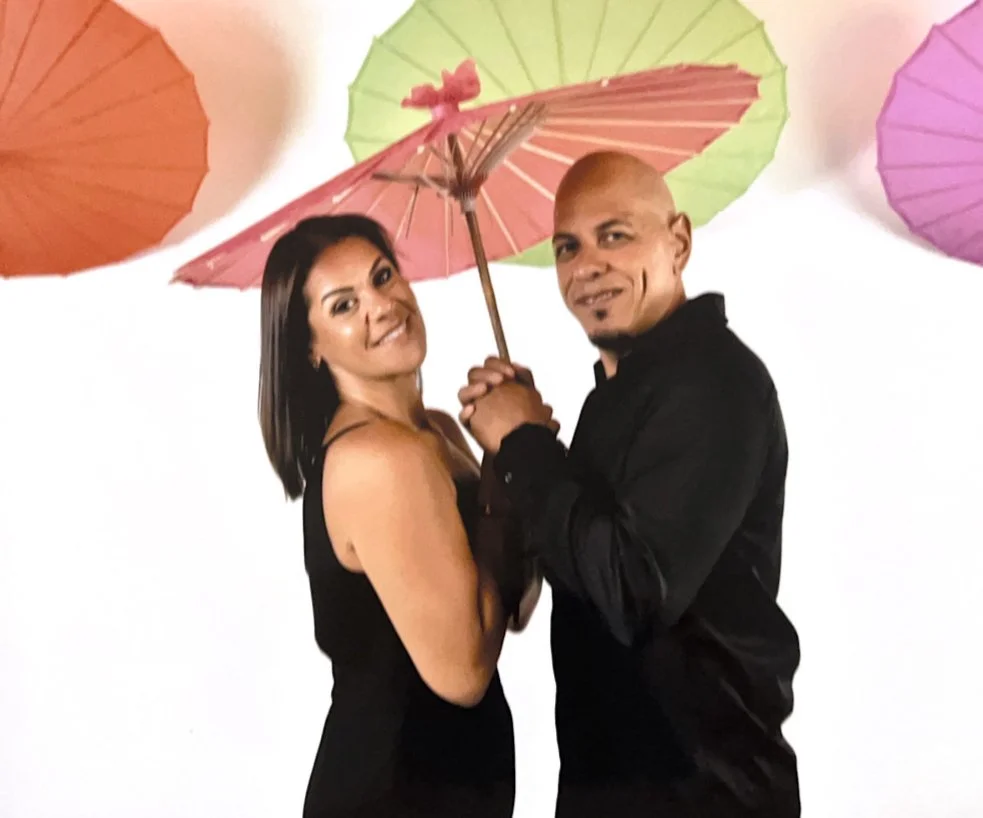 A woman and a man posing together with colorful umbrellas hanging overhead as a backdrop.