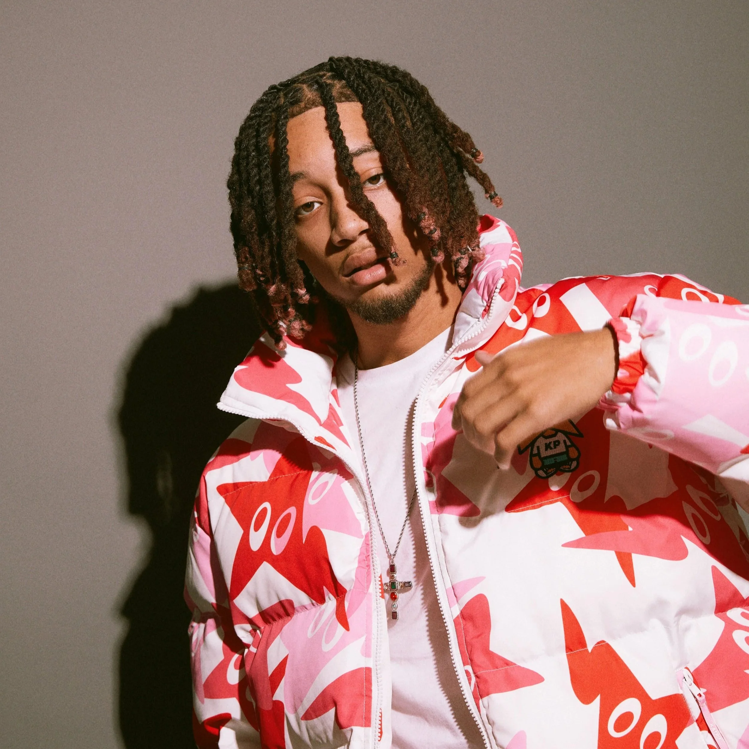 A young man with dreadlocks wearing a pink and white patterned jacket, looking at the camera against a plain gray background.