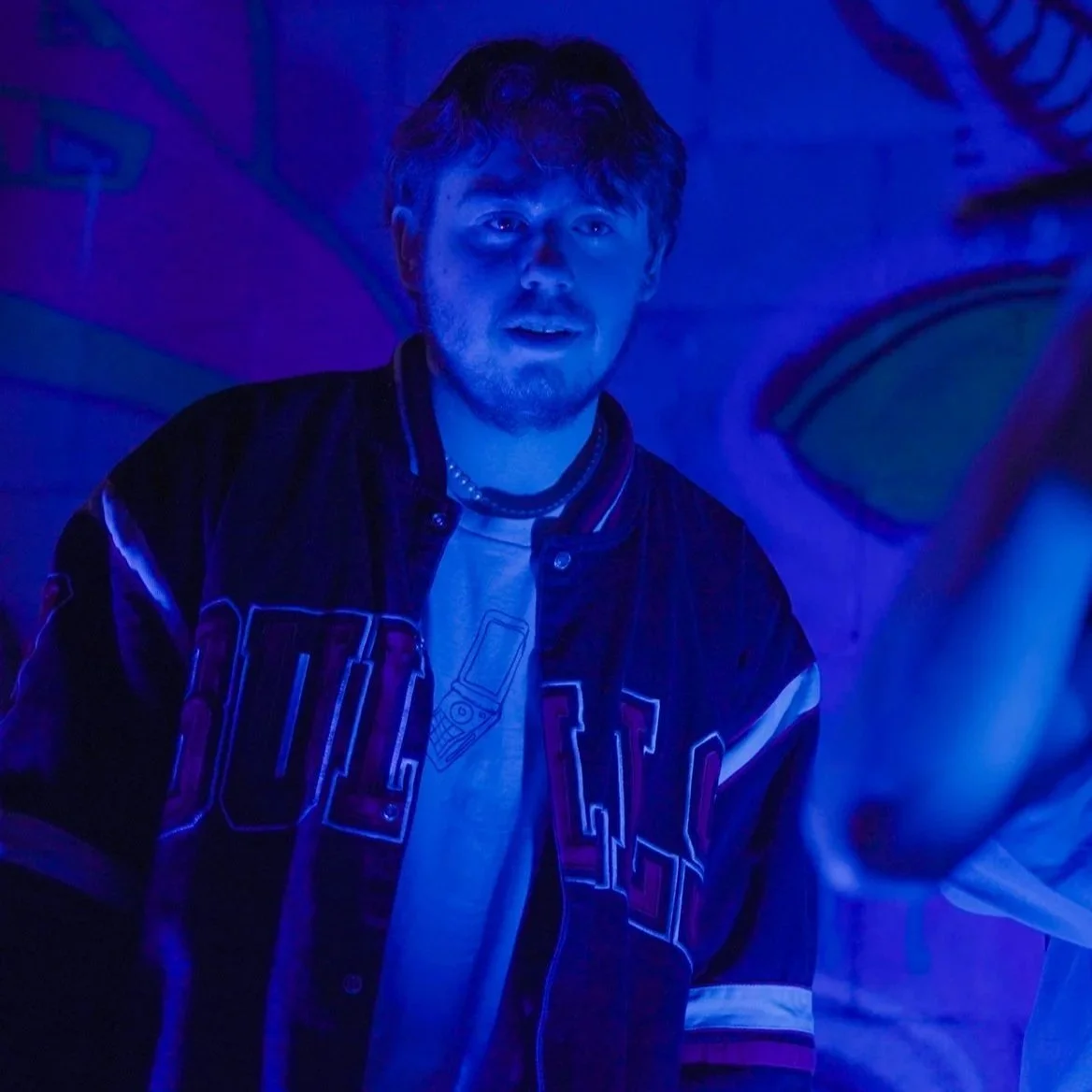 A young man with a beard and styled hair in a dimly lit room with blue light. He is wearing a black varsity jacket with white and red details and a white T-shirt, standing in front of a colorful mural.