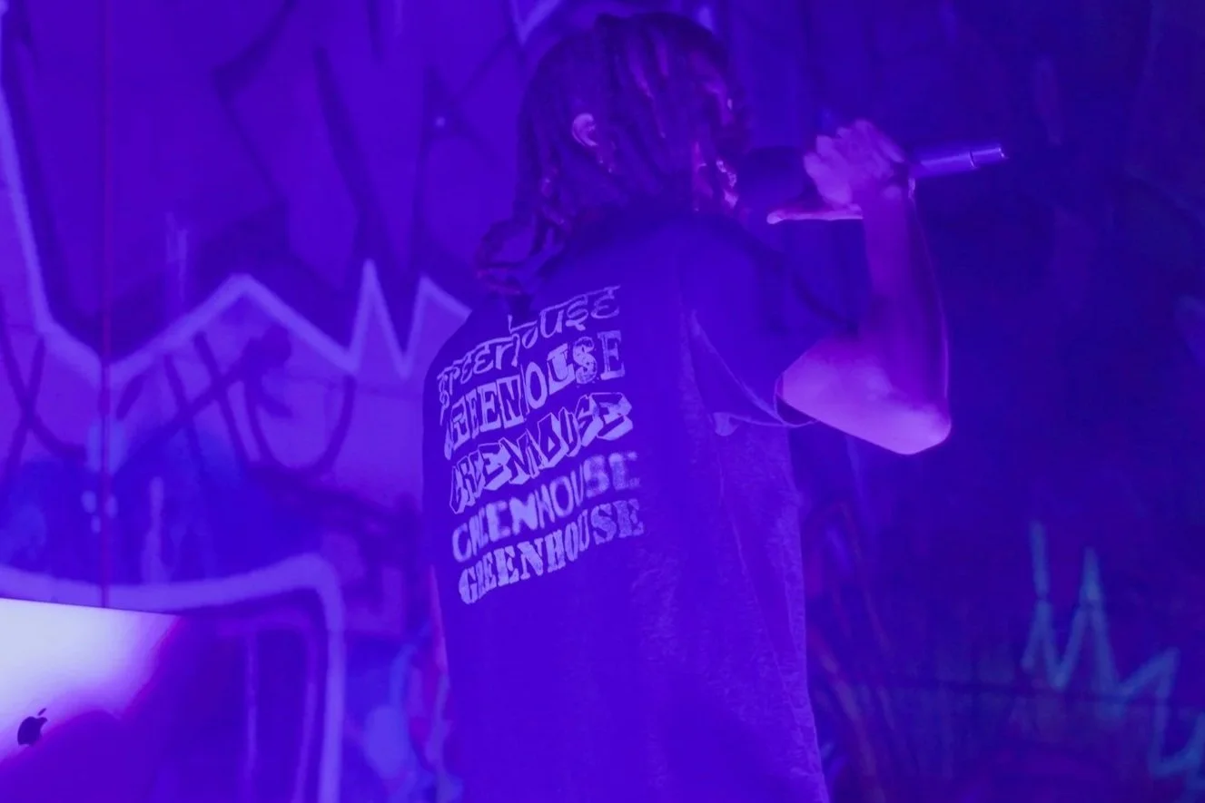 Person with dreadlocks singing into a microphone in a dimly lit environment with graffiti art on the wall behind them.