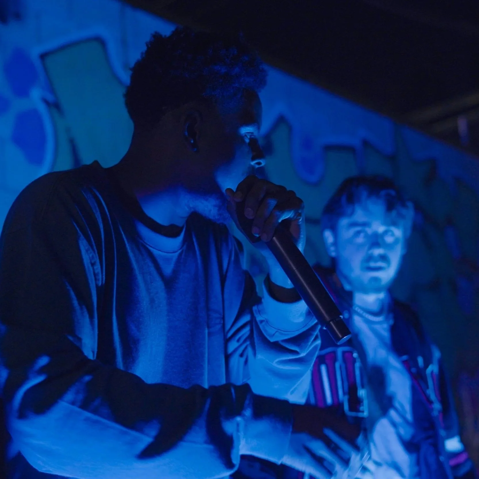A person with short curly hair, wearing a jacket, is holding a microphone, and standing in front of a wall with graffiti art. Another person with glasses and a striped shirt is in the background. The scene is lit with blue lighting.