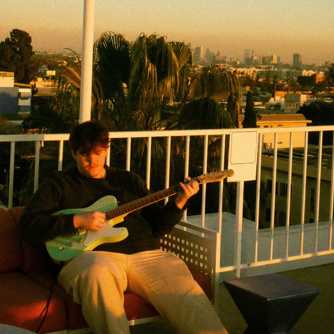 A young man with dark hair and glasses sitting on a patio, playing a green and white electric guitar, with a city skyline in the background during sunset.