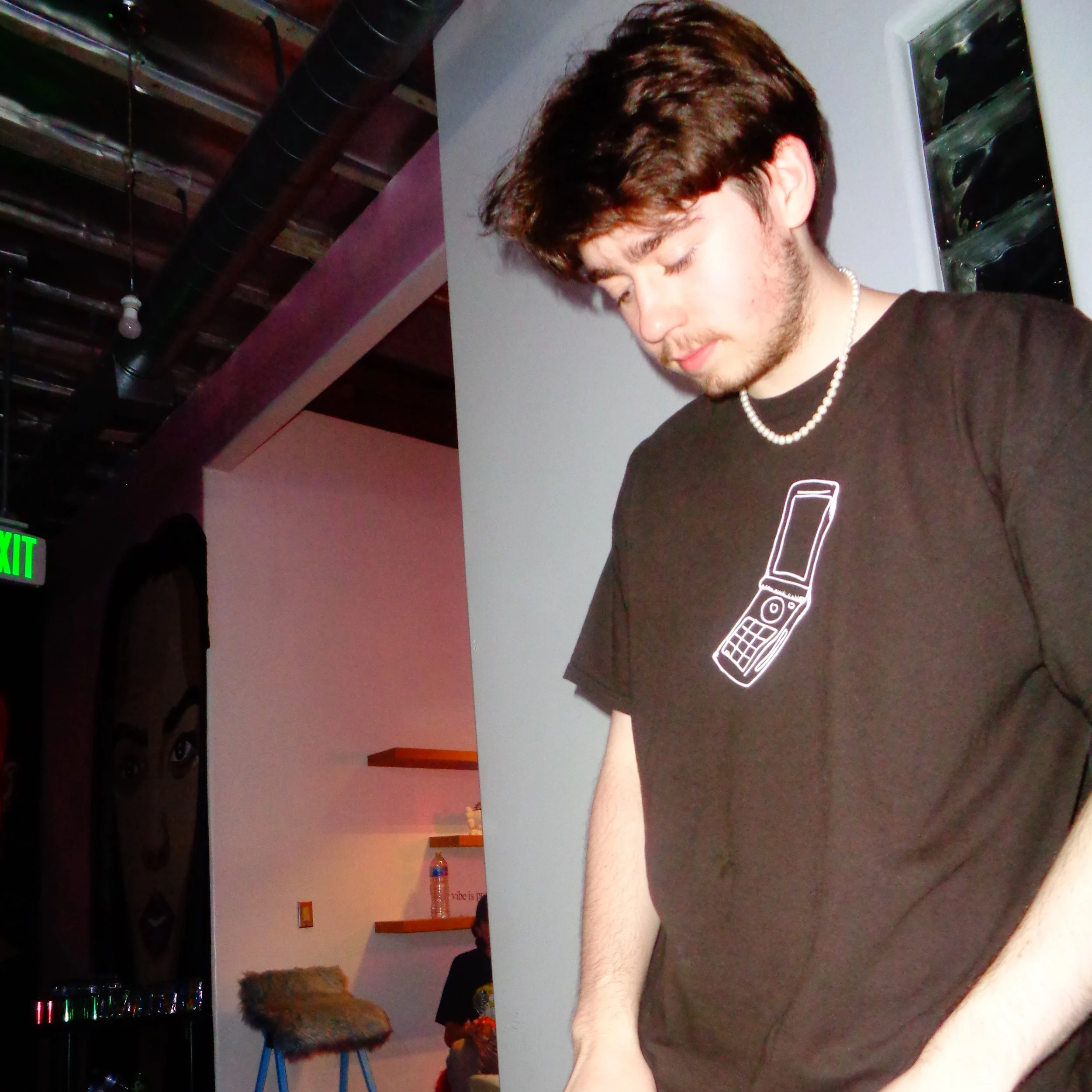A young man with brown hair and a beard wearing a black t-shirt and a pearl necklace, leaning over a white table or counter, with a bottle of water and a soda bottle in the background. The photo appears to be taken inside a bar or nightclub with visible ceiling pipes and a digital LED sign.
