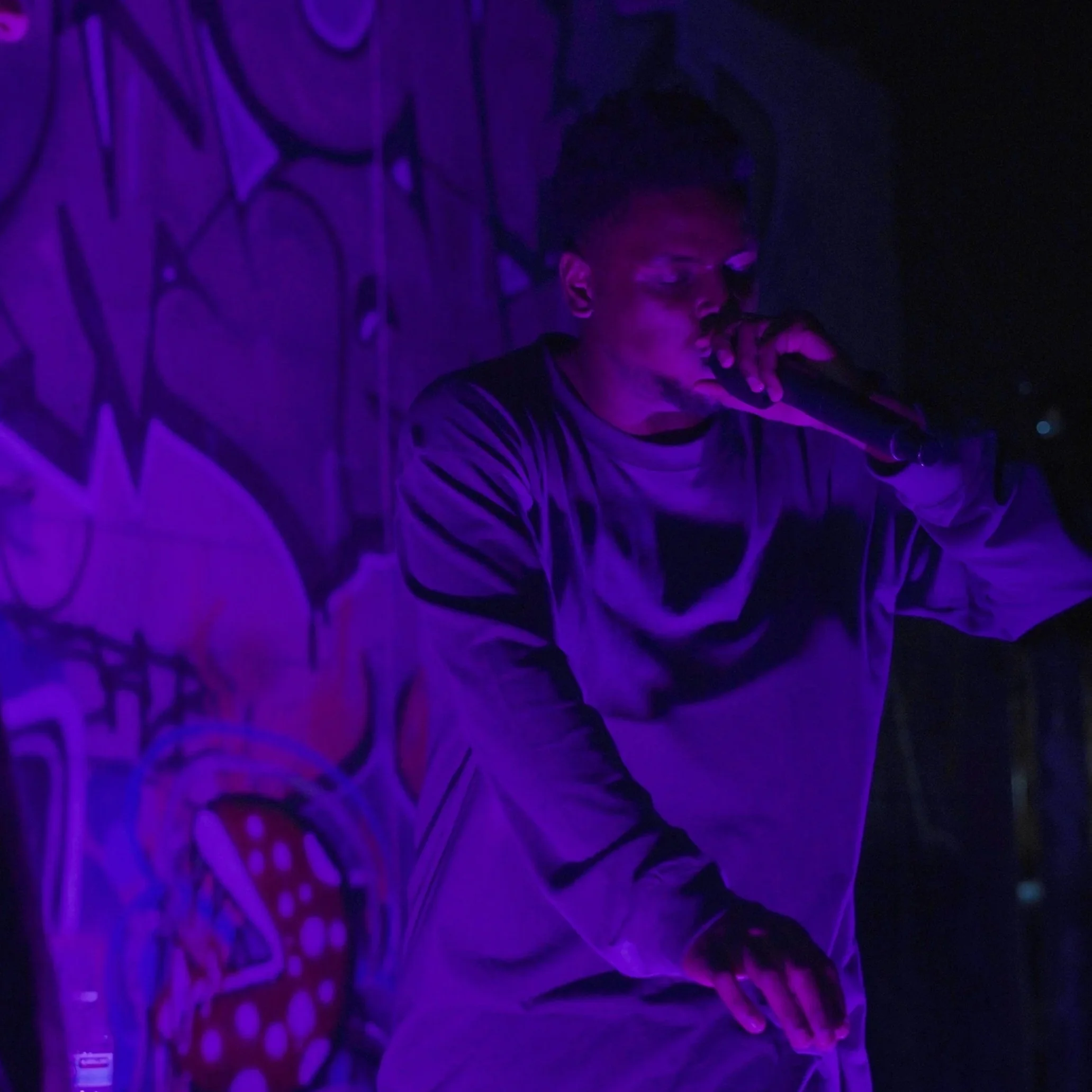 A person with short curly hair and glasses wearing a striped shirt, sitting in a dark room illuminated by purple and blue lighting, with graffiti art on the wall behind them.