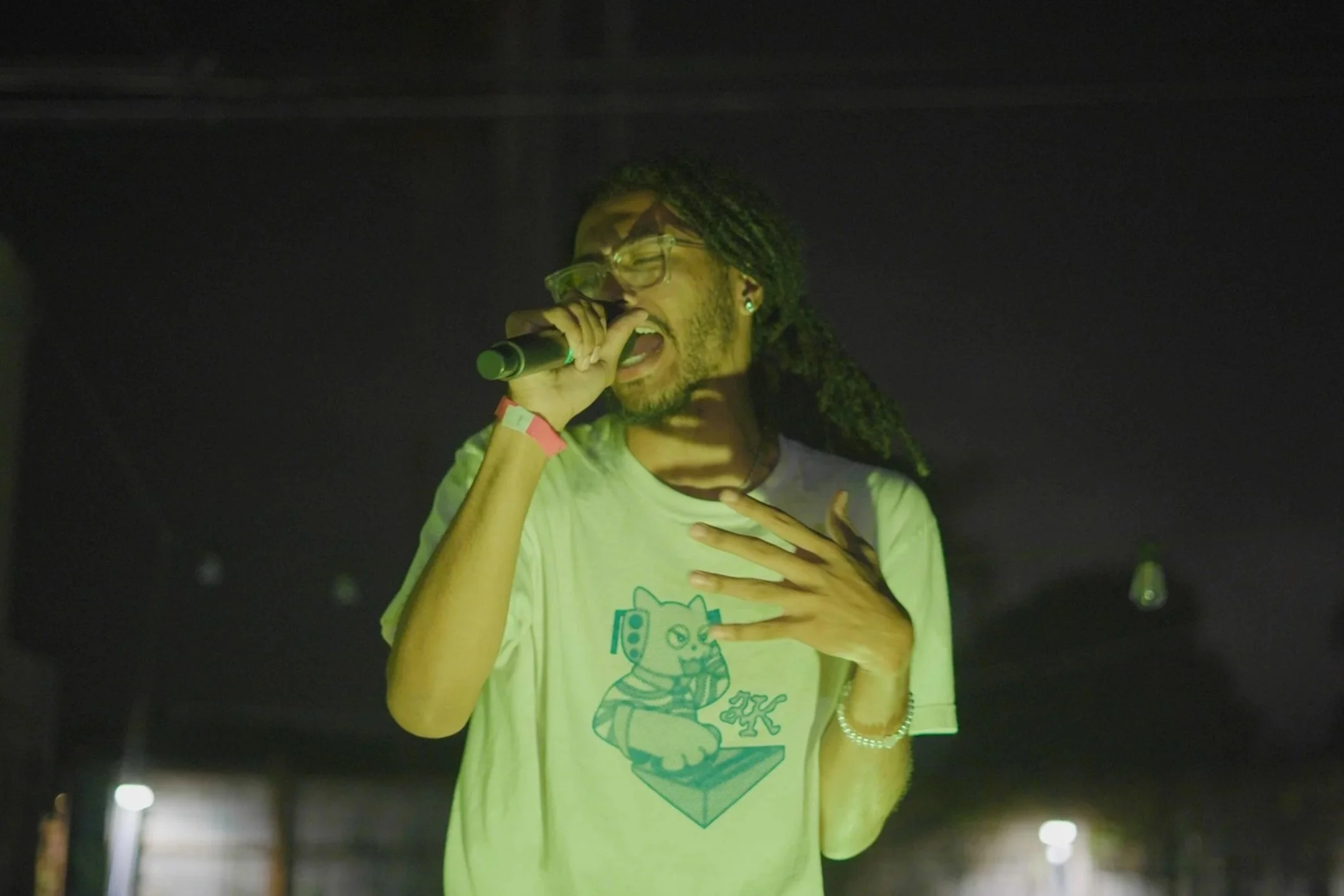 A person wearing glasses and a white T-shirt singing into a microphone on stage, with hand on chest, dark background, and dim, colorful stage lighting.