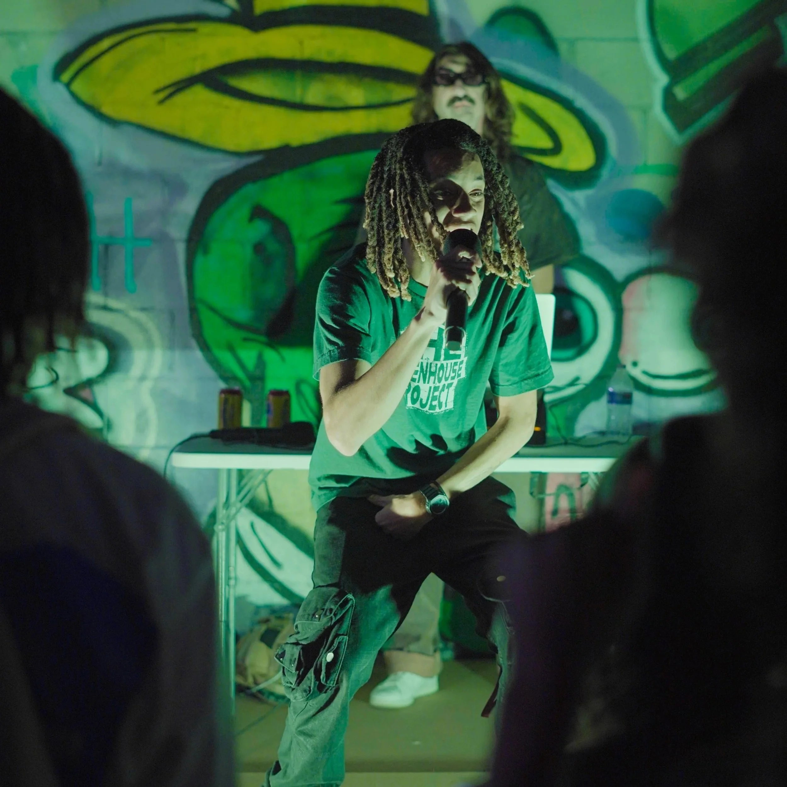 A person with dreadlocks holding a microphone, standing in front of a graffiti-covered wall with a large painted eye and face, illuminated by green lighting.