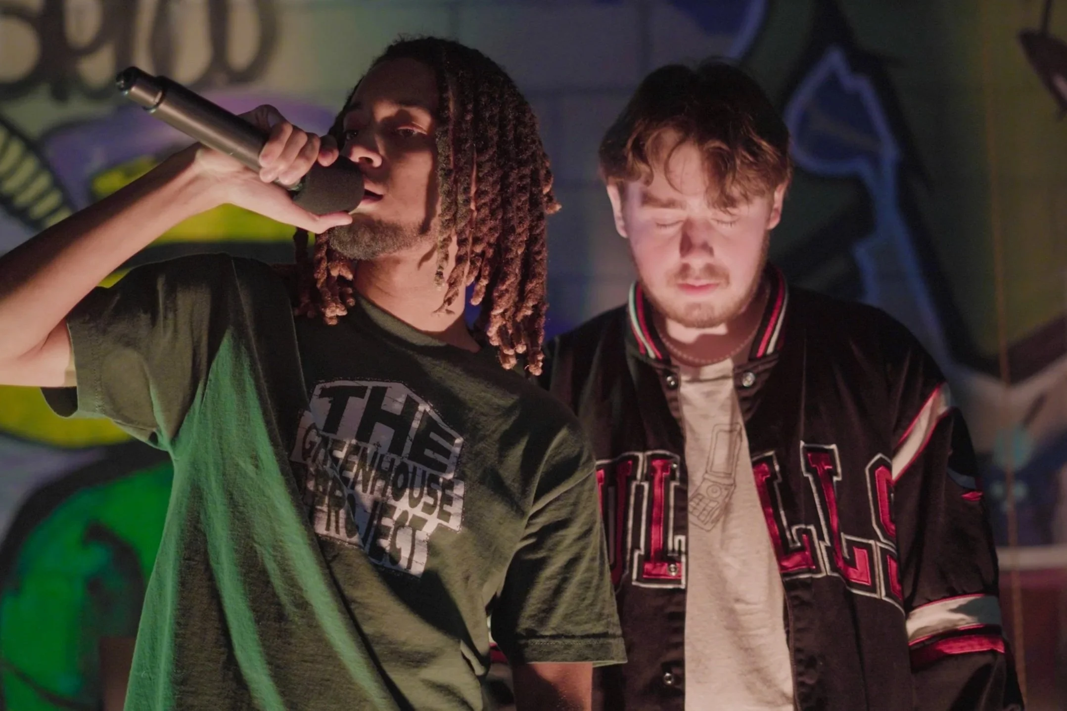 Two young men, one with dreadlocks and the other with short curly hair, are performing on stage with a microphone in front of him. They are under colorful stage lighting, with one wearing a graphic t-shirt and the other a varsity jacket.