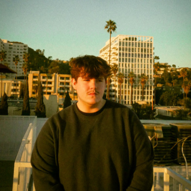 A young man standing outdoors with a cityscape and palm trees in the background during sunset.