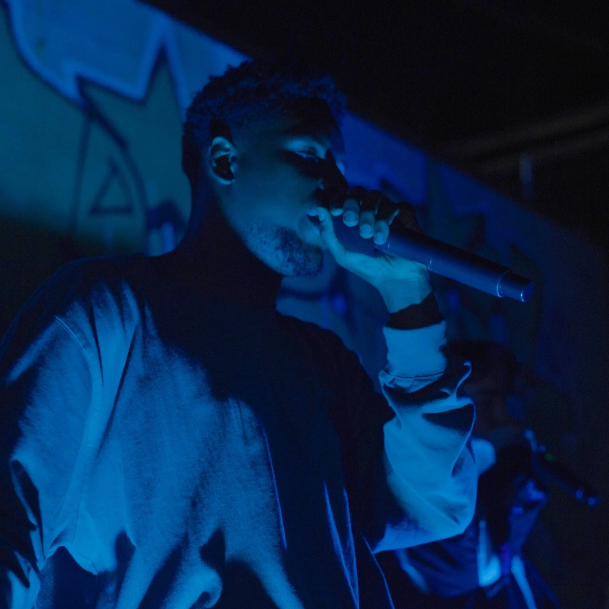 A man with glasses and curly hair is smiling and covering his mouth with his hand in a dimly lit, blue-toned environment.