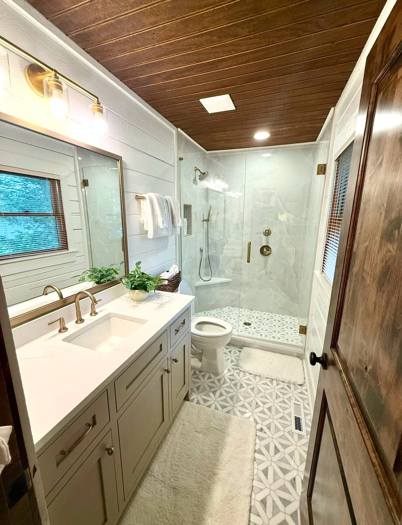 Hill Home Construction, Atlanta, Georgia. Modern bathroom with white vanity, marble countertop, a plant, large mirror, wooden ceiling, walk-in shower with glass door, patterned tile floor, window with blinds, toilet, and beige rugs.