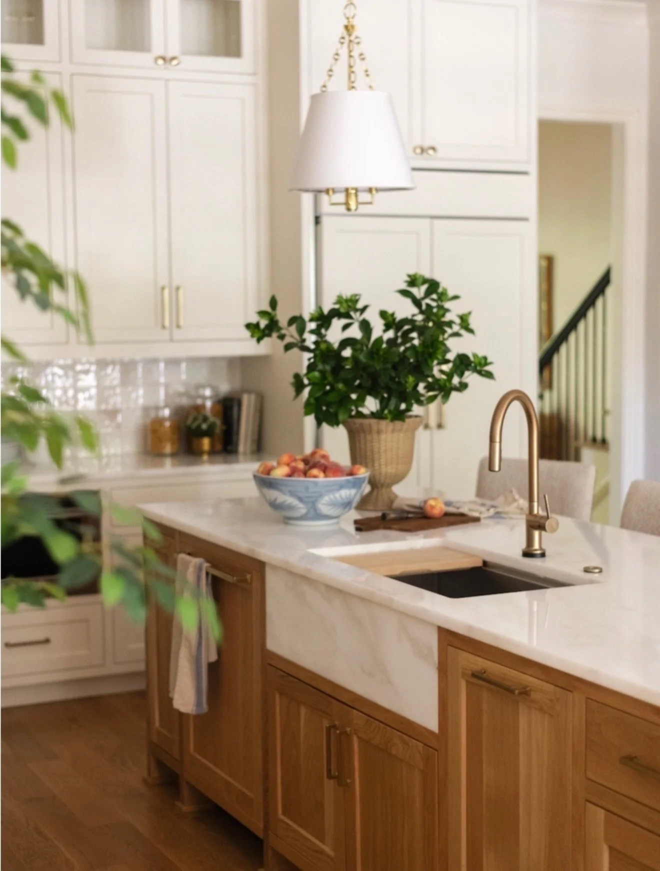 A kitchen with white upper cabinets, a marble island with a sink, a gold faucet, a large potted plant, a bowl of peaches, and a white hanging lamp.