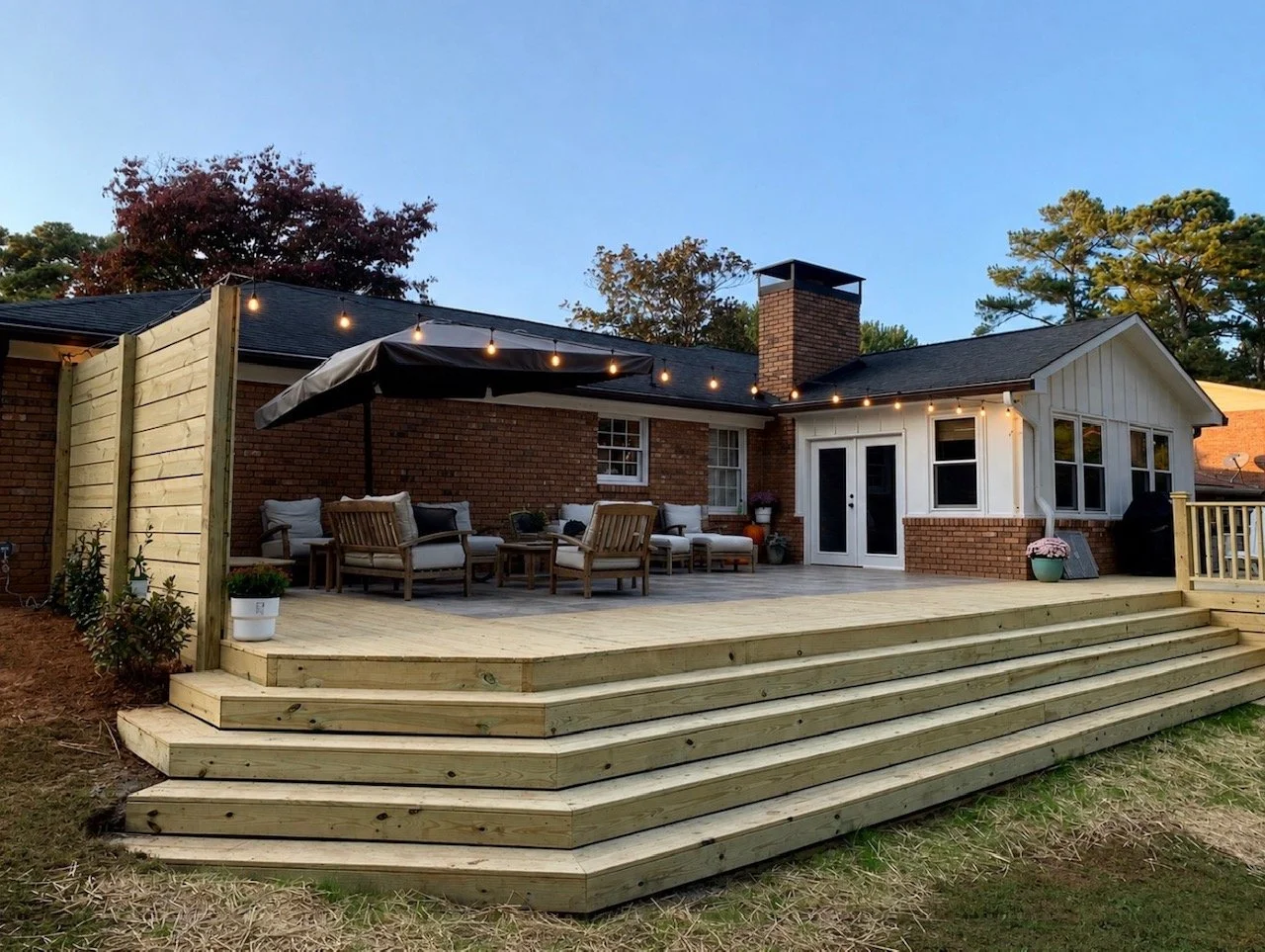 Backyard deck with seating area, string lights, a large umbrella, potted plants, and stairs leading down to grass, attached to a brick house with white siding and multiple windows.