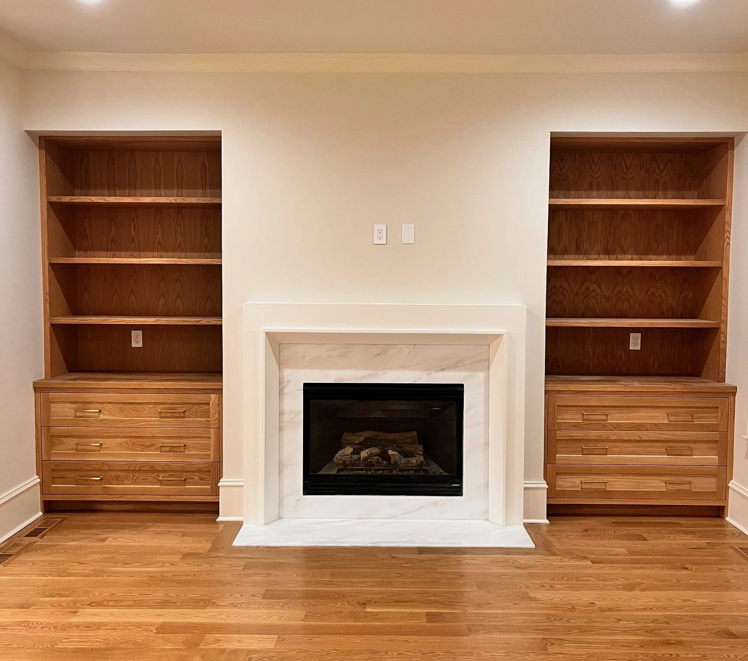 Empty built-in wooden bookshelves surrounding a white fireplace with a marble mantel in a living room.
