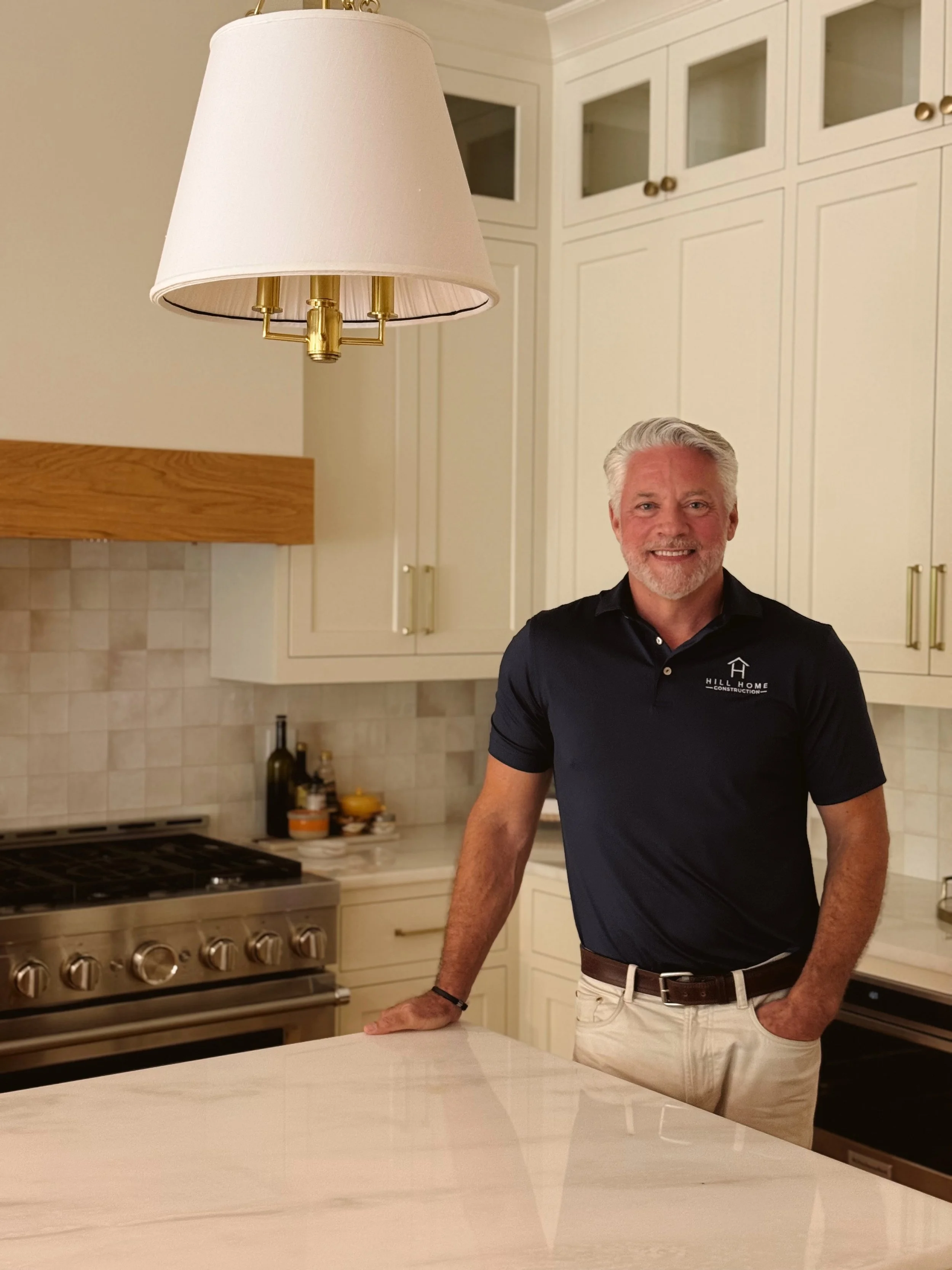 A smiling man with gray hair and a beard standing in a modern kitchen, wearing a black polo shirt and khaki pants. There is a large white lamp hanging from the ceiling, cream-colored cabinets, a stainless steel stove, and a white countertop.