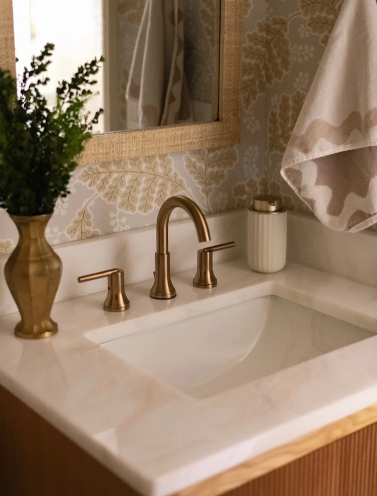 A bathroom countertop with a gold faucet, a potted plant in a gold vase, and a container with gold accents. A mirror reflects part of the countertop and a window with a curtain. The wall behind the counter has patterned wallpaper.