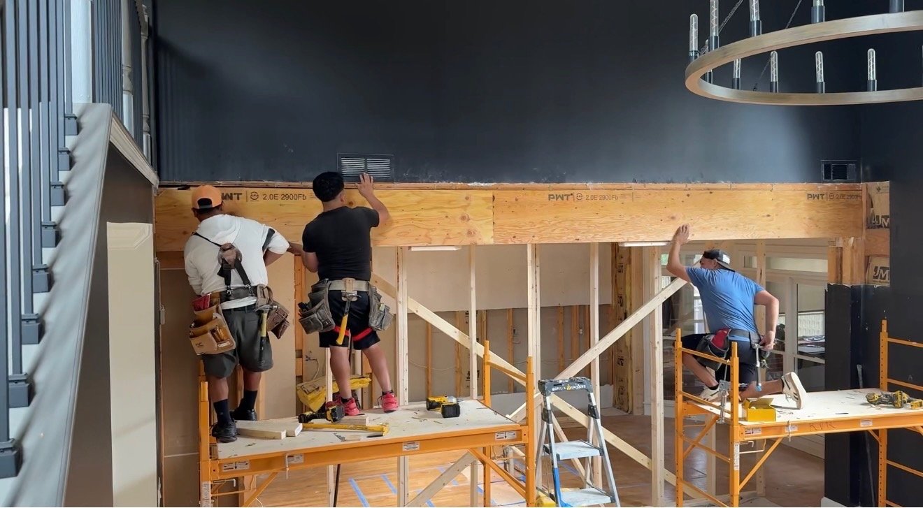 Hill Home Construction Project Construction Process. Three construction workers building a wall inside a building, working on wooden framing with tools around them.