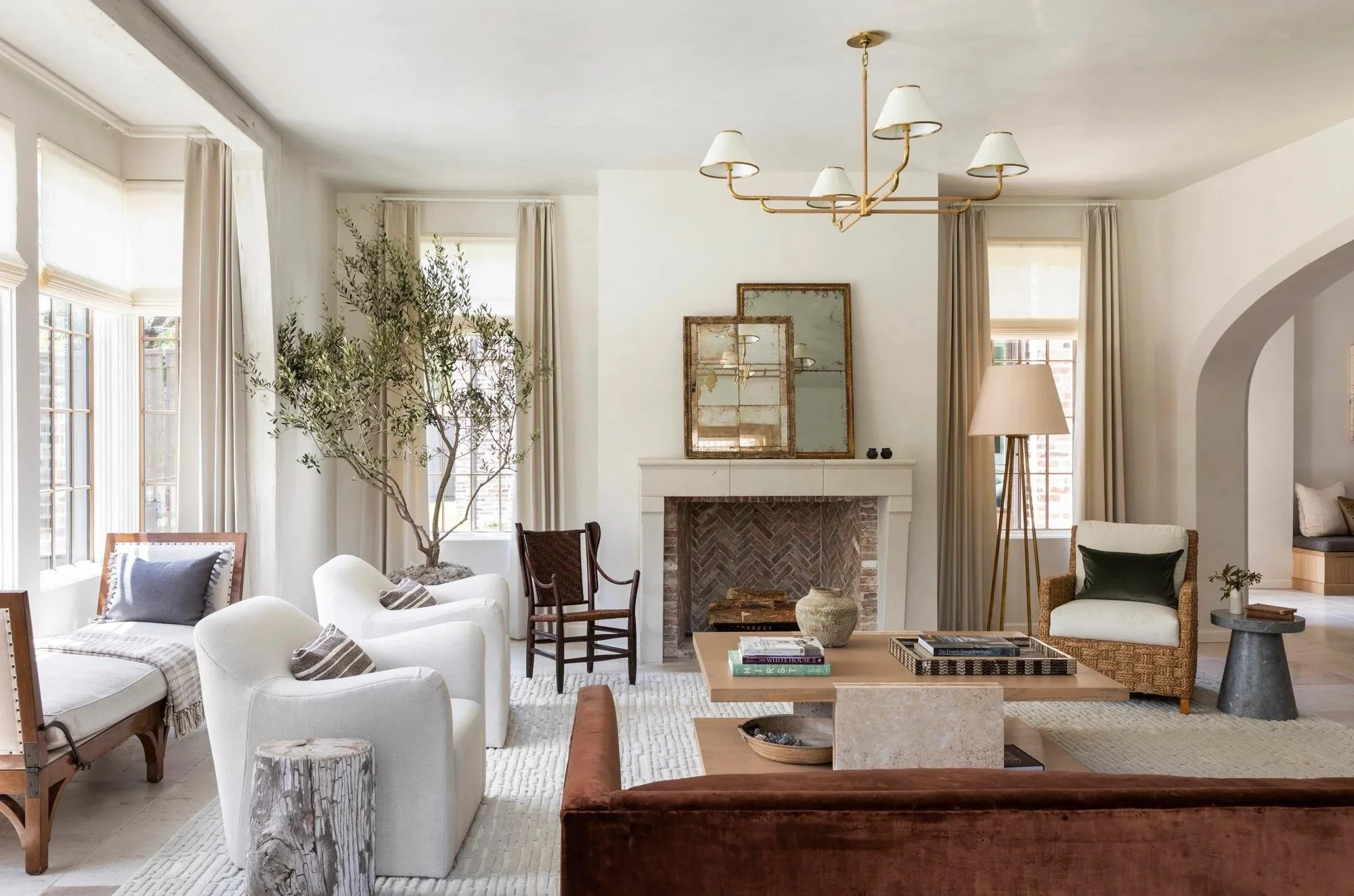 Living room with cream-colored walls, large windows with beige curtains, a fireplace with a mirror above, and various chairs and sofas arranged around a coffee table.