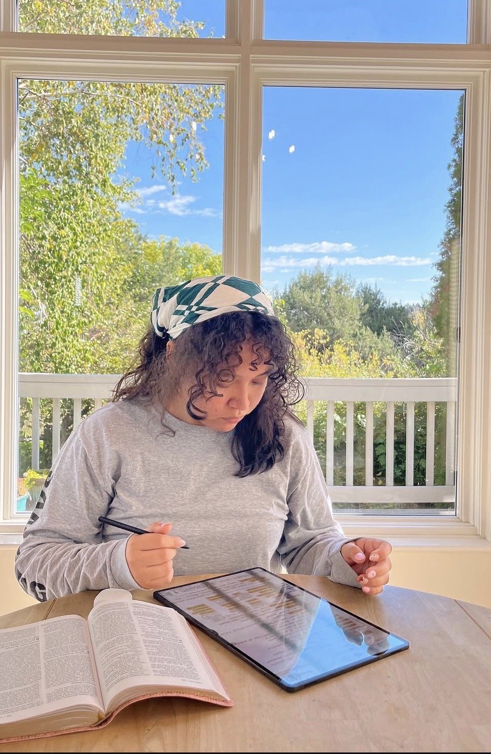 Woman sitting at a table in front of large window, working on a digital tablet with an open book nearby, on a sunny day with green trees outside.