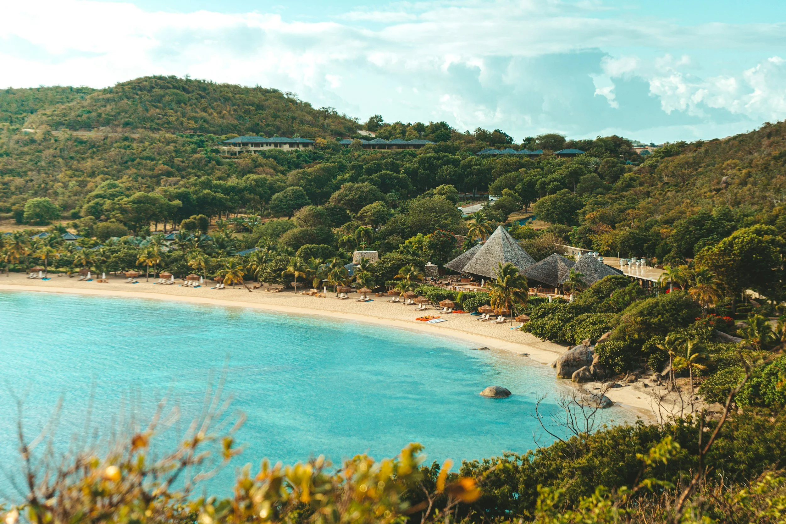A tropical beach with turquoise water, white sandy shore, palm trees, and thatched-roof huts, set against lush green hills and a partly cloudy sky.