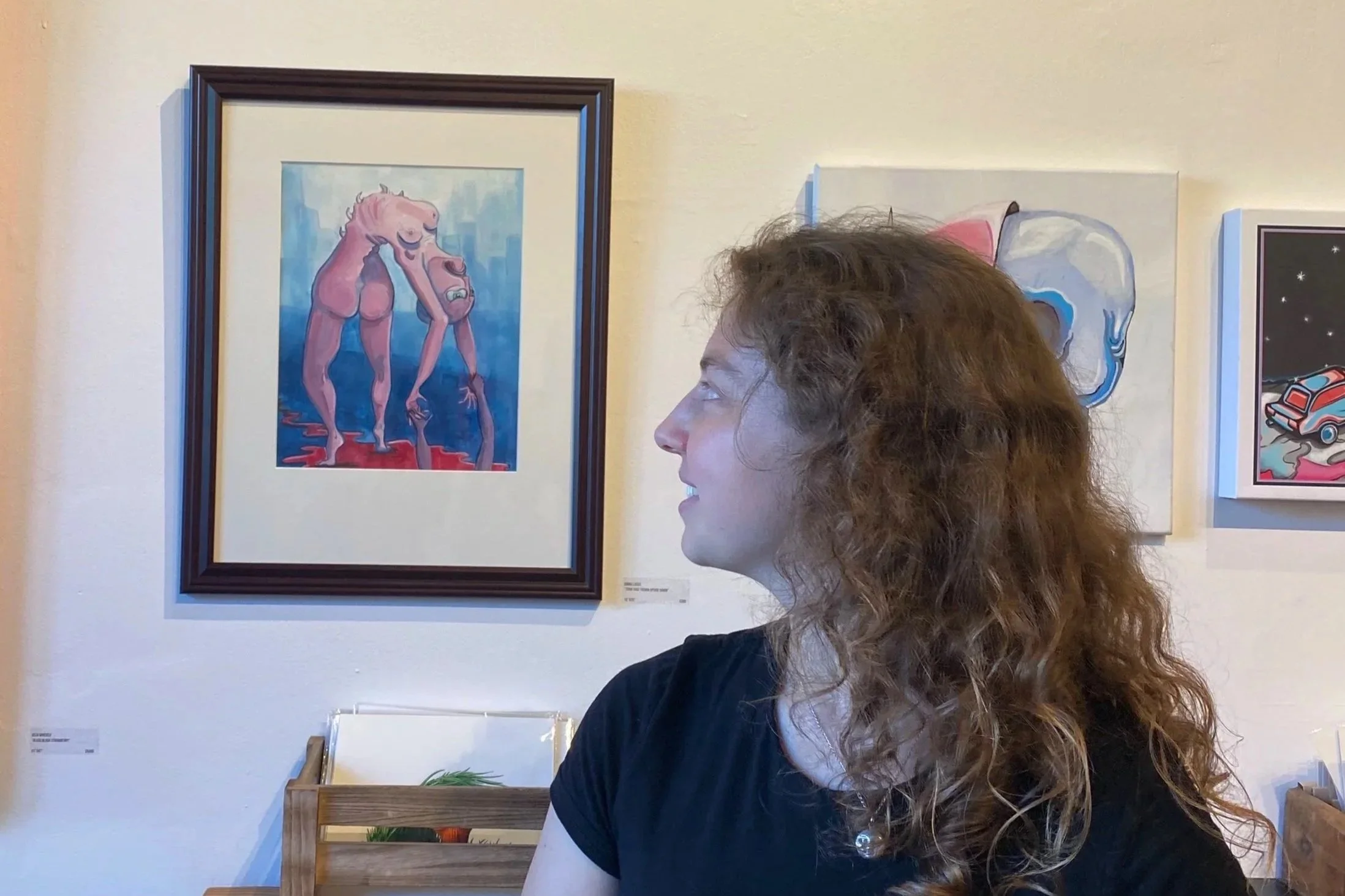 Emma smiling as she looks at artwork displayed on a gallery wall, including a painting of a pink humanoid figure in a blue and white background
