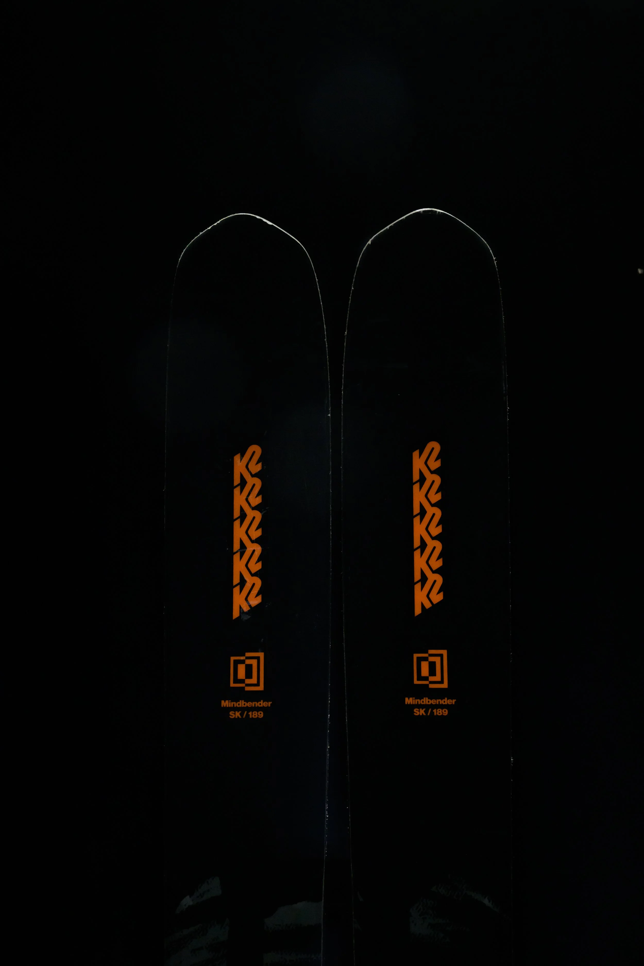 Two black skis with orange designs and the words 'Mindbender SK / 189' printed on them. The skis are set vertically against a dark background.