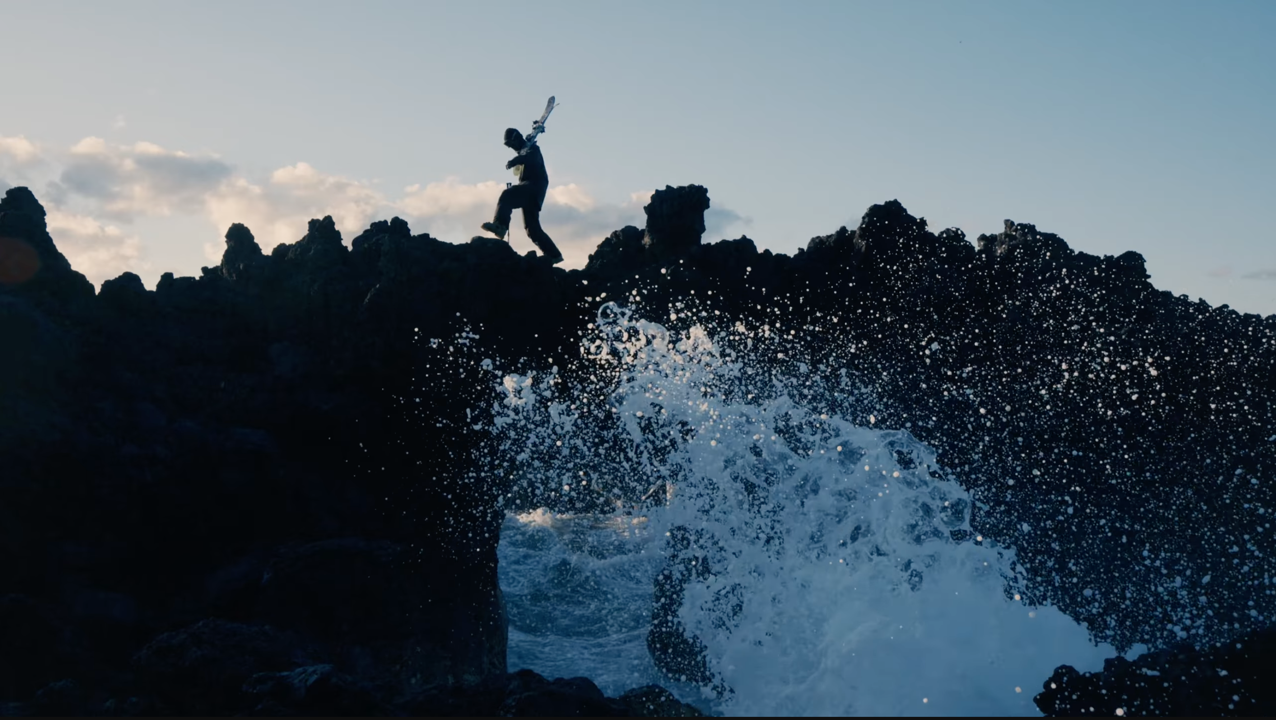 A person with a backpack and a helmet jumping from rocky cliffs into the ocean water as waves crash below, with a cloudy sky in the background.