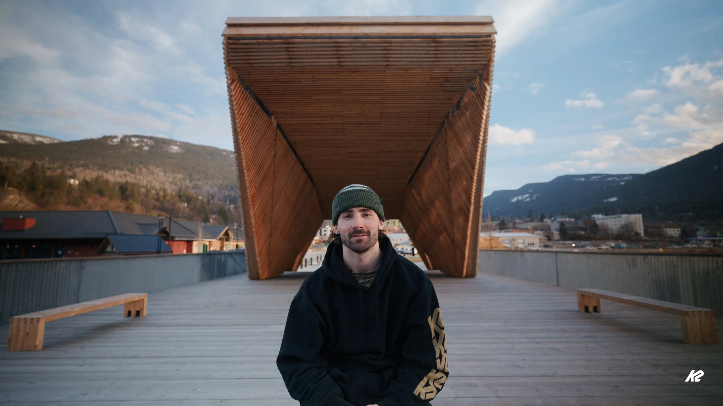 A man with a dark hoodie and green beanie standing on a wooden deck under a large wooden structure, with mountains and clouds in the background.