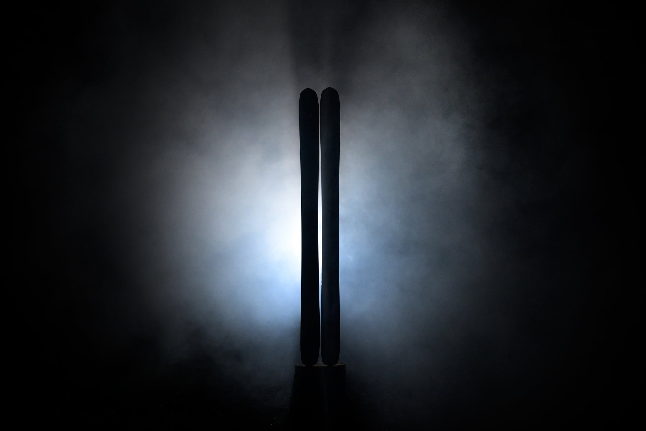 Two skis standing upright in a dark setting, illuminated from behind creating a silhouette with a diffused light glow.