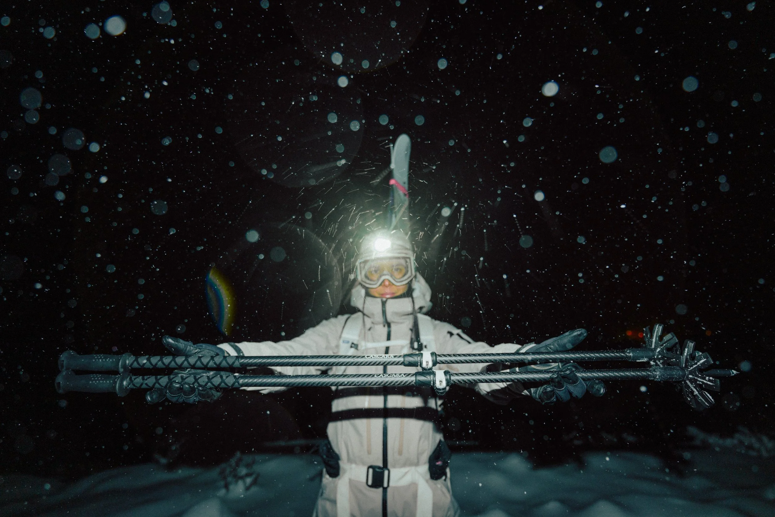 Person in winter gear with ski poles, wearing ski goggles and a helmet, standing in the snow at night with snowflakes falling around, illuminated by a bright light behind them.