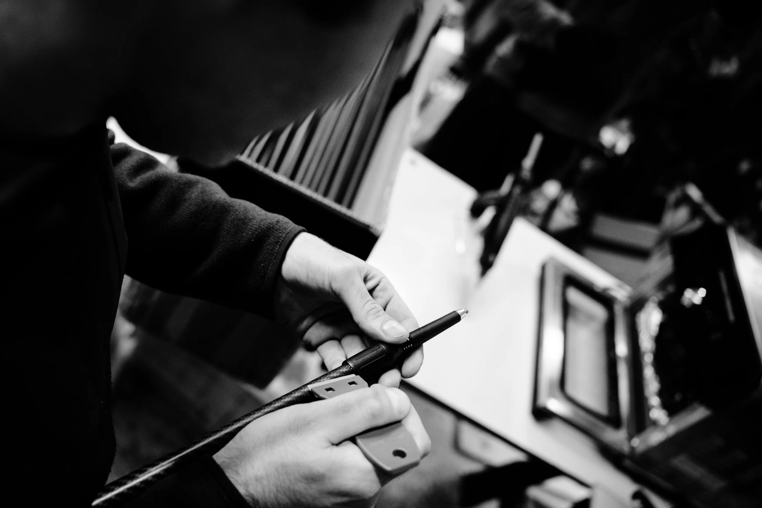 Close-up of a person's hands holding a pen and a piece of paper, with a keyboard and a stack of books or files in the background, all in black and white.