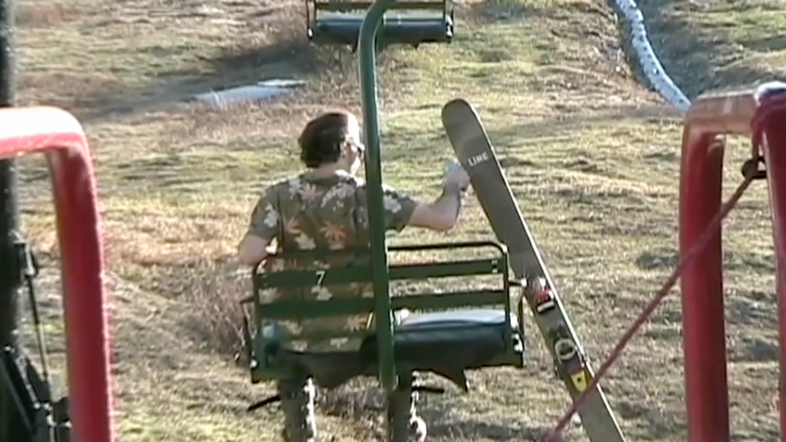 A person wearing a floral shirt sitting on a park bench in an outdoor playground area with grass and playground equipment nearby.