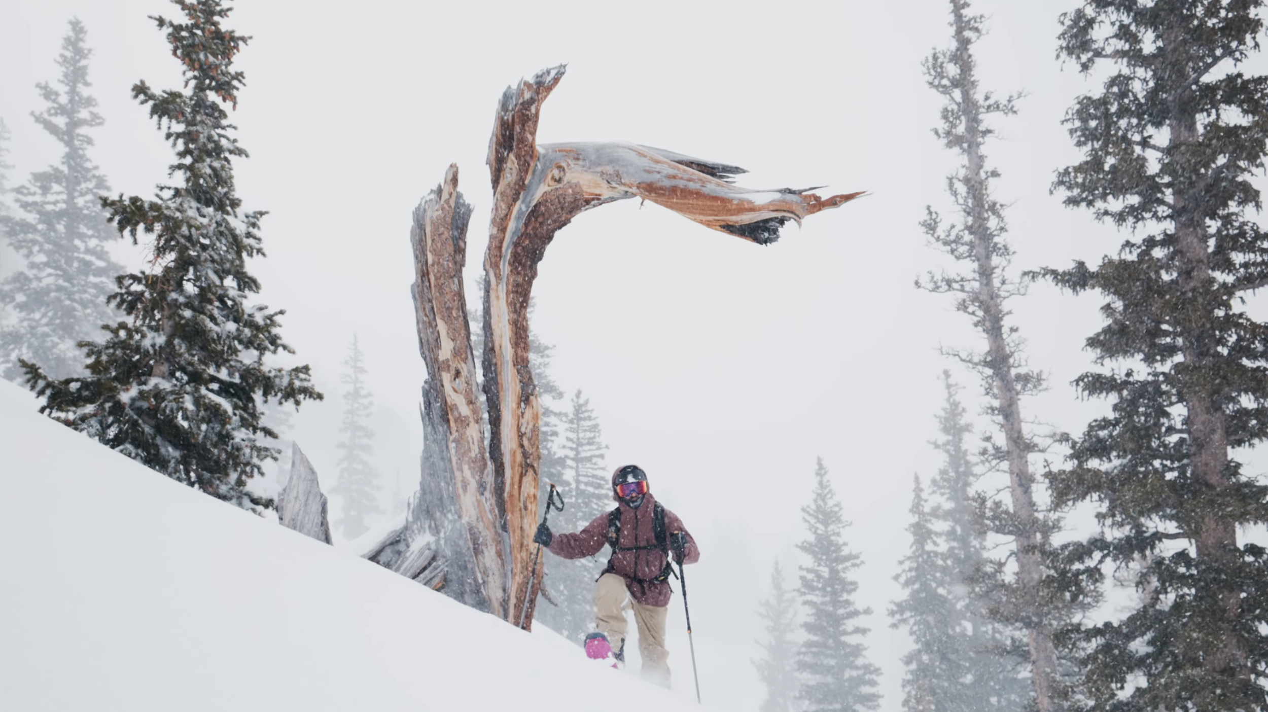 A person dressed in winter gear, including a red jacket, beige pants, gloves, and a helmet with goggles, is skiing down a snowy slope with tall pine trees and a misty sky in the background.