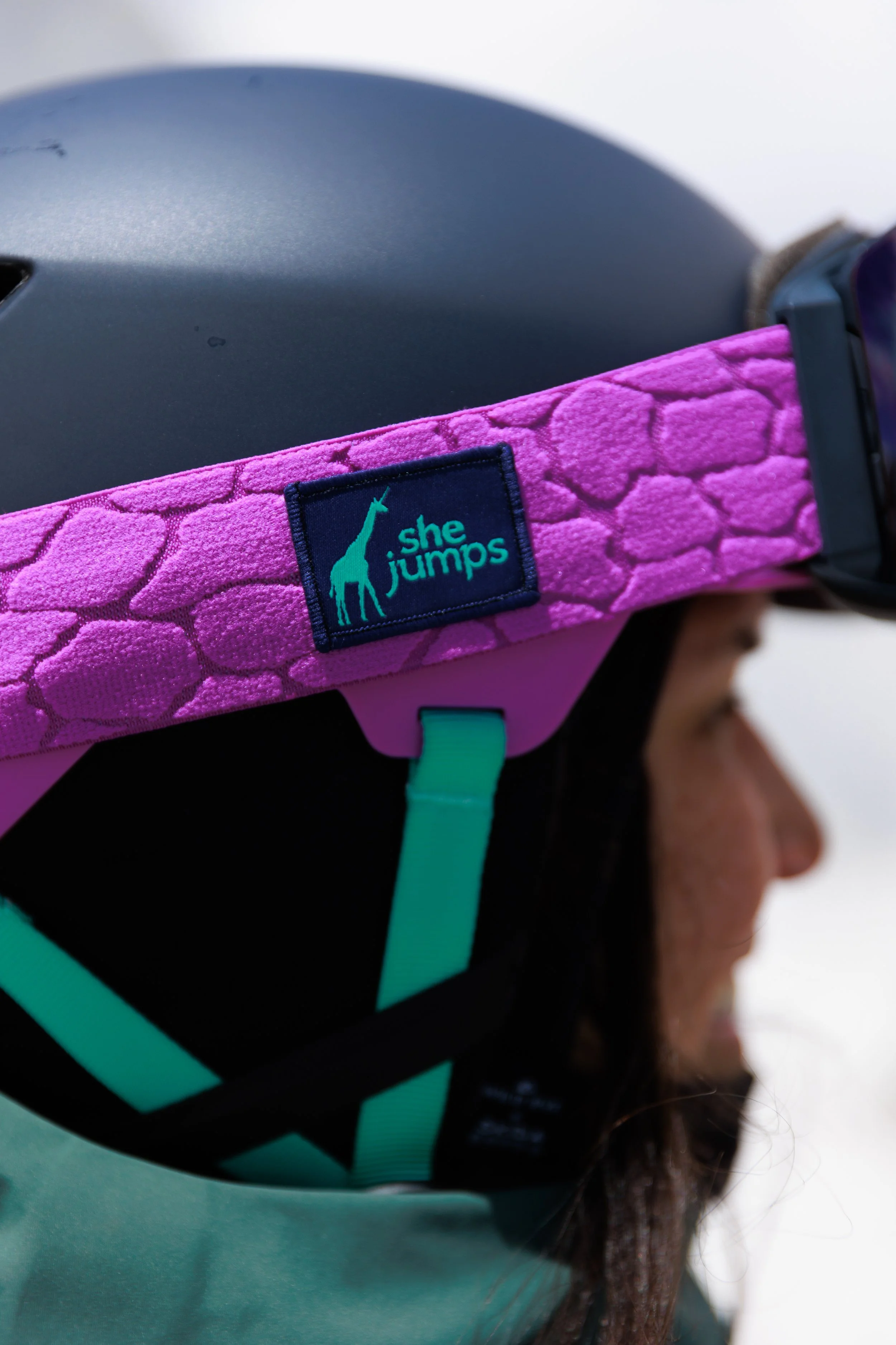 Person wearing a pink harness with a label that says 'she jumps' and a giraffe illustration, with a helmet on, facing sideways.